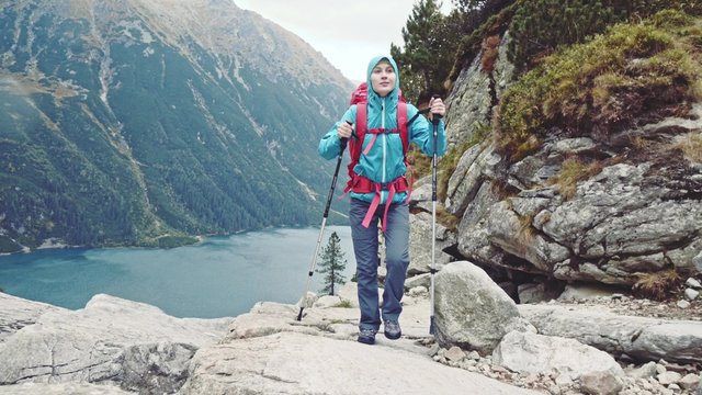 Hiker woman walking away from the cliff with the mountain lake view landscape behind her. Stabilized, 4K Ultra HD. Epic Steadicam hiking in a stormy wind, healthy lifestyle. Misty Mountains Series. 
