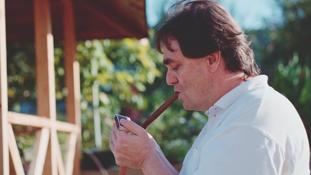 Adult Man is smoking cigar outdoors in the garden. Slow Motion 240 fps. Man is resting in a park during summer afternoon. Smoke in Slow Motion. 