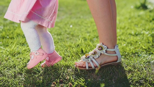 Little baby learns to walk. Slow Motion 240 fps. Mother is teaching her child to do the first steps on a green grass in summer. Close up on feet. Happy parenthood and childhood concept.