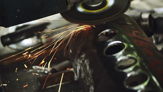 Worker grinding steel pipe, man using grinder to work on piece of metal in workshop, grinding sparks flying around.