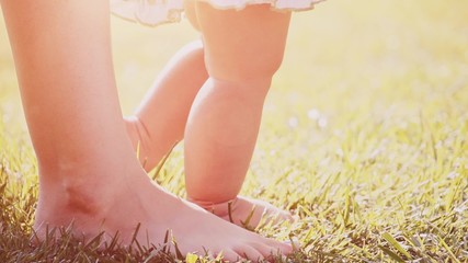 Little baby learns to walk. Close up on feet. Slow Motion 240 fps. Mother is teaching her child to do the first steps on a green grass in summer. Toddler is learning to walk outdoors on a green lawn.