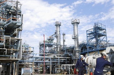 two oil-workers with pump machinery, refinery in background