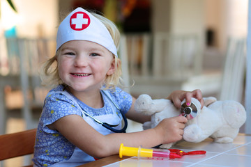 Funny blonde toddler girl playing doctor role game examinating her puppy using stethoscope sitting at small white table in playroom at home, school or kindergarten