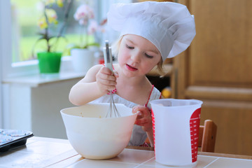 Baking with children: little happy kid, adorable toddler girl in white chef hat mixing dough...