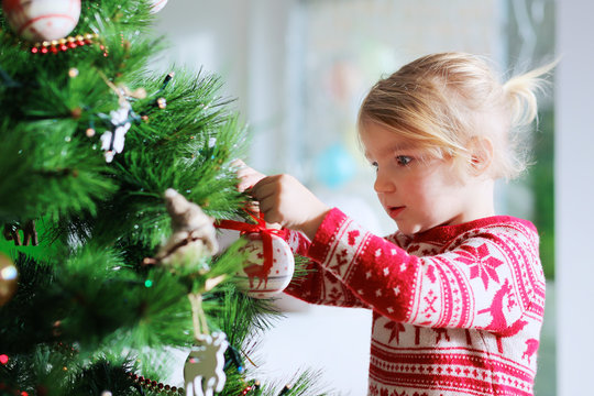 Little girl decorating christmas tree with toys and baubles. Cute kid preparing home for xmas celebration.