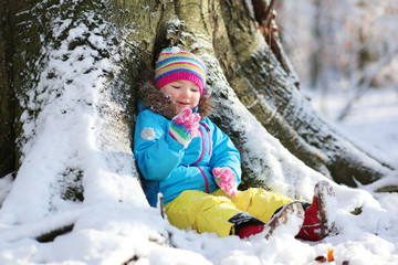 Fototapeta na wymiar Active healthy toddler girl wearing colorful snowsuit and bright knitted hat and mittens enjoying snow playing in a beautiful snowy forest on sunny winter day