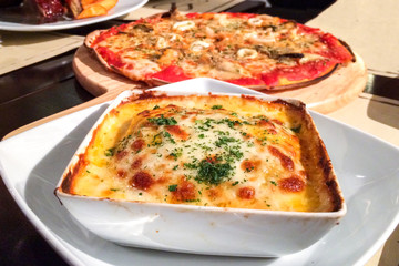 Traditional Italian Lasagna made with Minced Beef Bolognese Sauce topped with Basil Leaves served on a White Plate with Blur Pizza in Background
