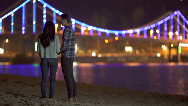 The couple (pair) sit on the beach, talk, kiss and hug by picturesque embankment, city and bridge lights background. Shot with Red Cinema Camera