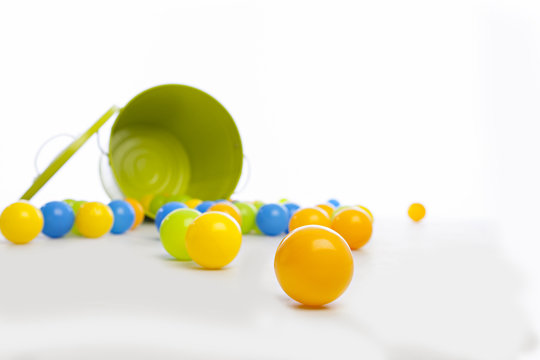 close up of yellow ball wiht green metal bucket that has spilled a large number of multi-colored balls on a white backdrop and floor
