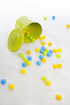 green metal bucket that has spilled a large number of multi-colored balls on a white backdrop and floor
