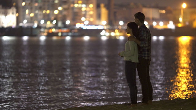 The couple (pair) stand on the beach hug and dream by city lights background. Shot with Red Cinema Camera