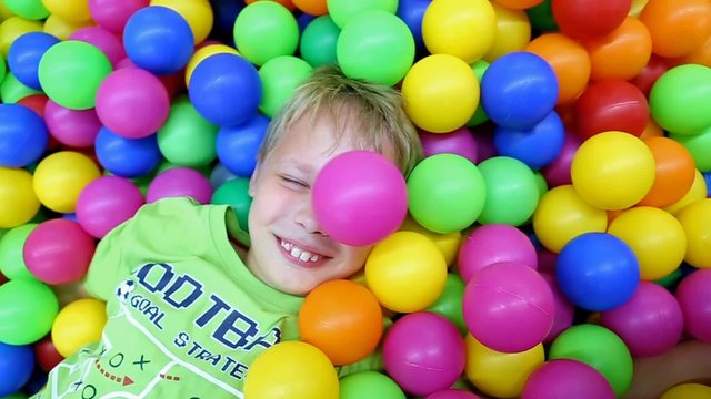 Cute kid laying among many colorful plastic toy balls at children playground. Little boy playing at ball pool. Slow motion video clip