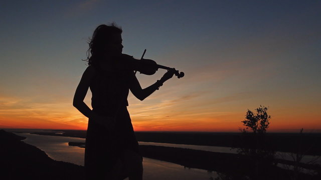 Young woman on the mountain playing the violin on river background at sunset.