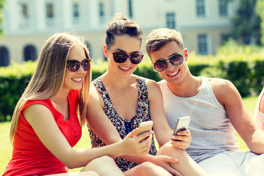 smiling friends with smartphones sitting in park