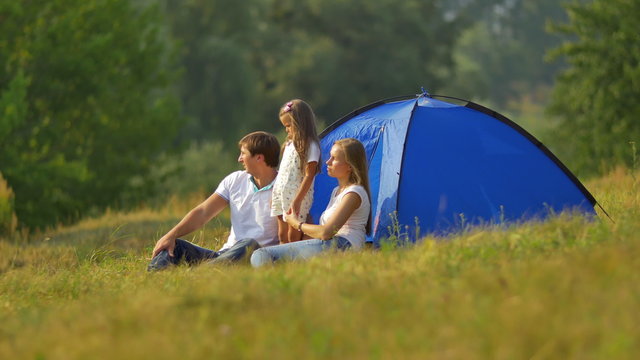 The family (father, mother and daughter) sit near tent on the hill by forest background. Real time capture. Shot with Red Cinema Camera