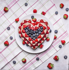 homemade cake In the form of heart  with blueberries and strawberries On a white plate on a background of a striped pink napkins with berries top view close up