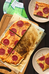 Homemade pepperoni or salami pizza cut in pieces on baking paper on wooden board and served on plates on the side, photographed overhead on dark wood with natural light