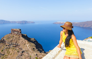 Attractive woman wearing hat and yellow dress enjoying the view of volcanic island in the early morning on Santorini, Mediterranean sea, Greece
