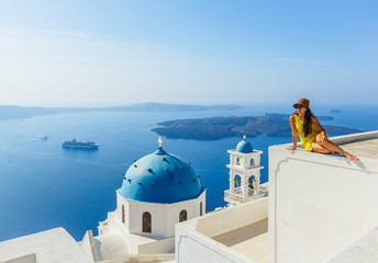 Female tourist in yellow dress, wearing hat enjoying view on Santorini in the morning on building above famous church, Mediterranean sea, Greece