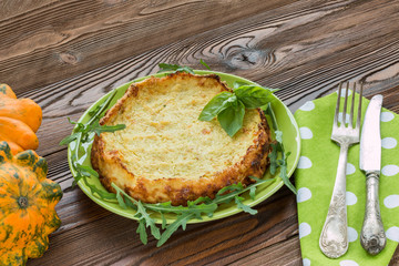 squash casserole decorated by basil leaf and arugula on light green plate at wooden table with squash fruits and light green napkins at white polka dots pattern and old silver cutlery