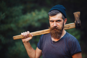 Brutal brunette bearded man in warm hat with a hatchet in the woods on a background of trees