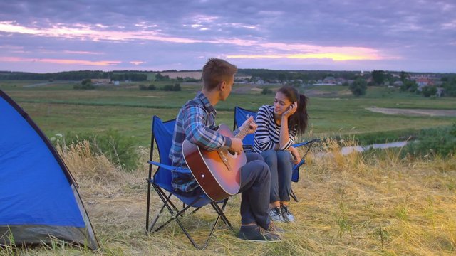Couple sit near bonfire and camping tent: man play the guitar, woman listen