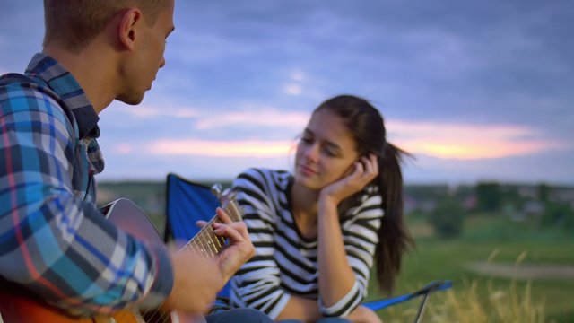 Couple sit near bonfire and camping tent: man play the guitar, woman drink tea