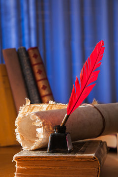 Red quill pen, black inkwell and a rolled papyrus sheet on an old book with a blue background