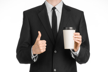 Business lunches coffee theme: businessman in a black suit holding a white blank paper cup of coffee with a brown plastic cap isolated on a white background in the studio, advertising coffee