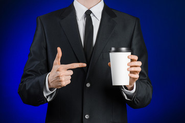 Business lunch and coffee theme: businessman in a black suit holding a white blank paper cup of coffee with a brown plastic cap on a dark blue background isolated in the studio, advertising coffee