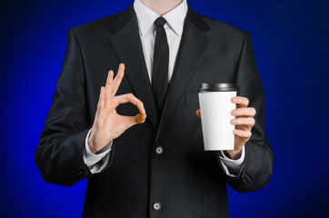 Business lunch and coffee theme: businessman in a black suit holding a white blank paper cup of coffee with a brown plastic cap on a dark blue background isolated in the studio, advertising coffee