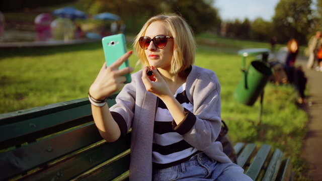 Girl sitting on the bench and improving her makeup while looking on smartphone
