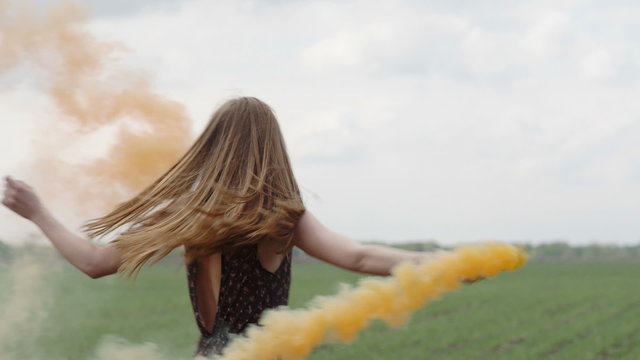 Happy young woman in dark dress with floral print having fun outdoor in geen field. Beauty girl with orange smoke bomb spinning and laughing. middle long shot slow motion