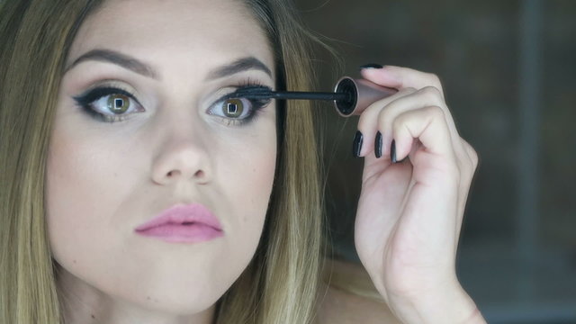 Pretty woman applying mascara to her eyelashes in front of bathroom mirror