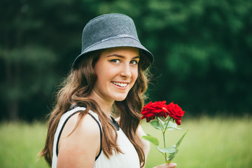 Beautiful smiled little girl with hat standing on a meadow, holding a rose and looking at camera....