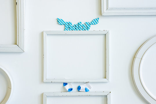 Empty frame for photograph, decorated with wooden letters "Baby" and little white and blue socks on the white wall.