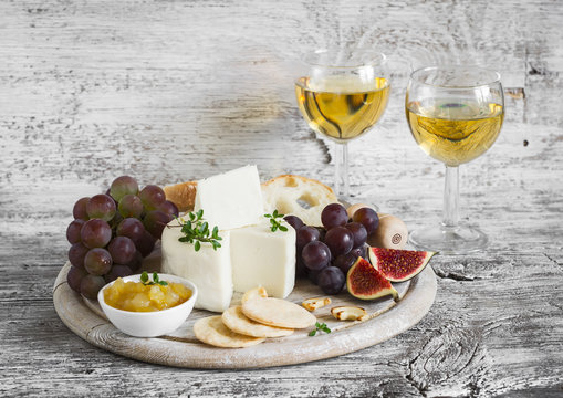 delicious appetizer to wine - ham, cheese, grapes, crackers, figs, nuts, jam, served on a light wooden board, and two glasses with white wine on bright wooden surface