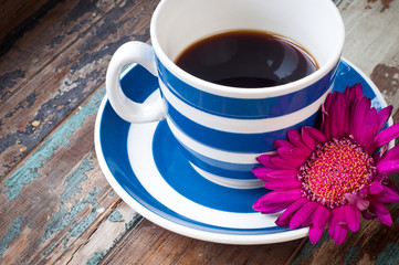 Freshly made coffee in a blue stripey cup served on a rustic wooden table with a wild flower.