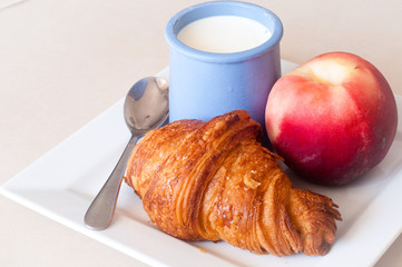 Healthy breakfast of freshly baked croissant and a peach with a serving of chilled yogurt in a blue ceramic pot.