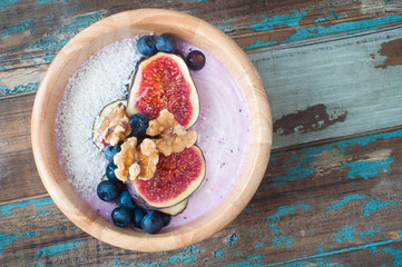 Smoothie bowl of blueberry and strawberry blended with kefir yogurt and topped with fresh fig, blueberries, walnuts and coconut. Served in a wooden bowl on a rustic wooden table.