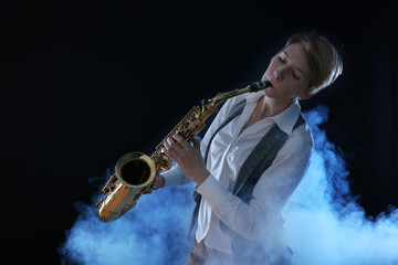 Attractive woman plays saxophone on dark background in the smoke