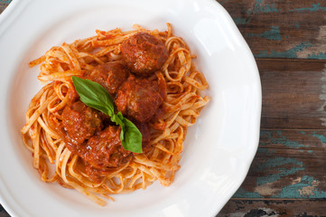 Spaghetti and meatballs in a tomato and herb sauce topped with fresh basil. A traditional italian pasta dish served in a white bowl on a rustic wooden table.