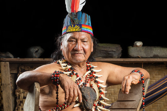 An elderly Ecuadorian Amazonian shaman in a colorful feather headdress and beaded necklaces sits adorned with bone pendants in a rural village, preserving the native culture of Amazonia.