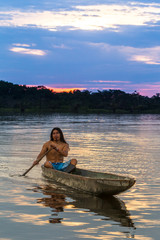 An indigenous man paddles a canoe on the Amazon River, surrounded by lush greenery in the isolated...