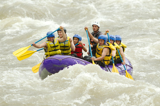A group of energetic people in a purple raft paddle through churning whitewater rapids in Ecuador, wearing yellow life vests and helmets, splashing through the green water on a team adventure.