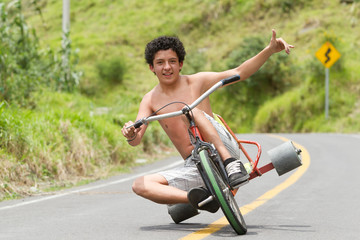 Obraz premium A smiling shirtless man rides a drift trike down a paved road in a green rural area with his arm raised in Ecuador, a carefree adventurous downhill ride.