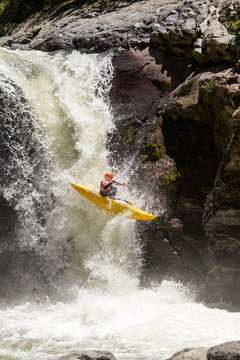 A kayaker in a bright yellow kayak descends a powerful waterfall surrounded by dark moss-covered rocks in the Sangay region of Ecuador, capturing the intensity of whitewater kayaking.