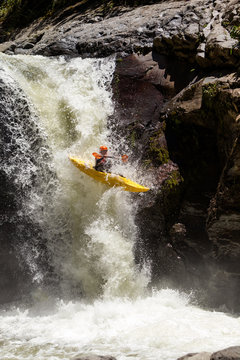 A kayaker in a bright yellow kayak navigates down a powerful waterfall surrounded by dark wet rocks in Banos, Ecuador, splashing water and dynamic motion capturing the thrill of whitewater kayaking.