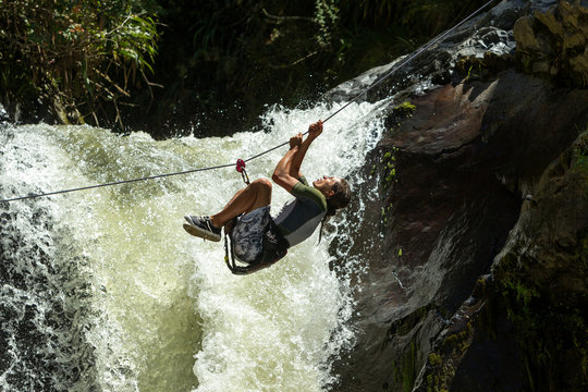A man hangs on a steel zipline cable soaring over a rushing waterfall surrounded by lush green jungle and mountains in Ecuador, an exhilarating extreme adventure on the canopy line.