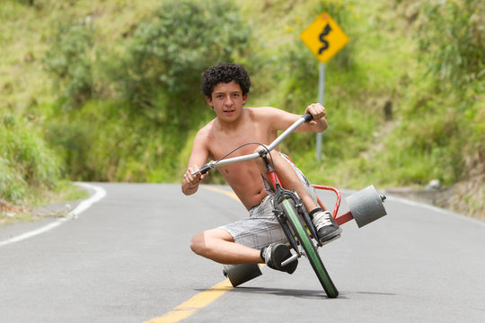 A shirtless young man rides a custom chopper-style drift trike down a curving mountain road in lush green rural Ecuador, leaning into the turn in a moment of motion and freedom.